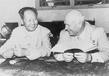 A balding man and a younger Chinese man sit and smile, the balding man holding a fan