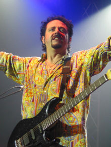 Lukather stands on stage with a black Ernie Ball Music Man "Luke" electric guitar and raises his arms to the crowd.