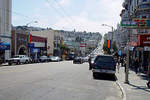 A street extending into the background. Stores line both sides of the street, and many light poles carry rainbow flags.